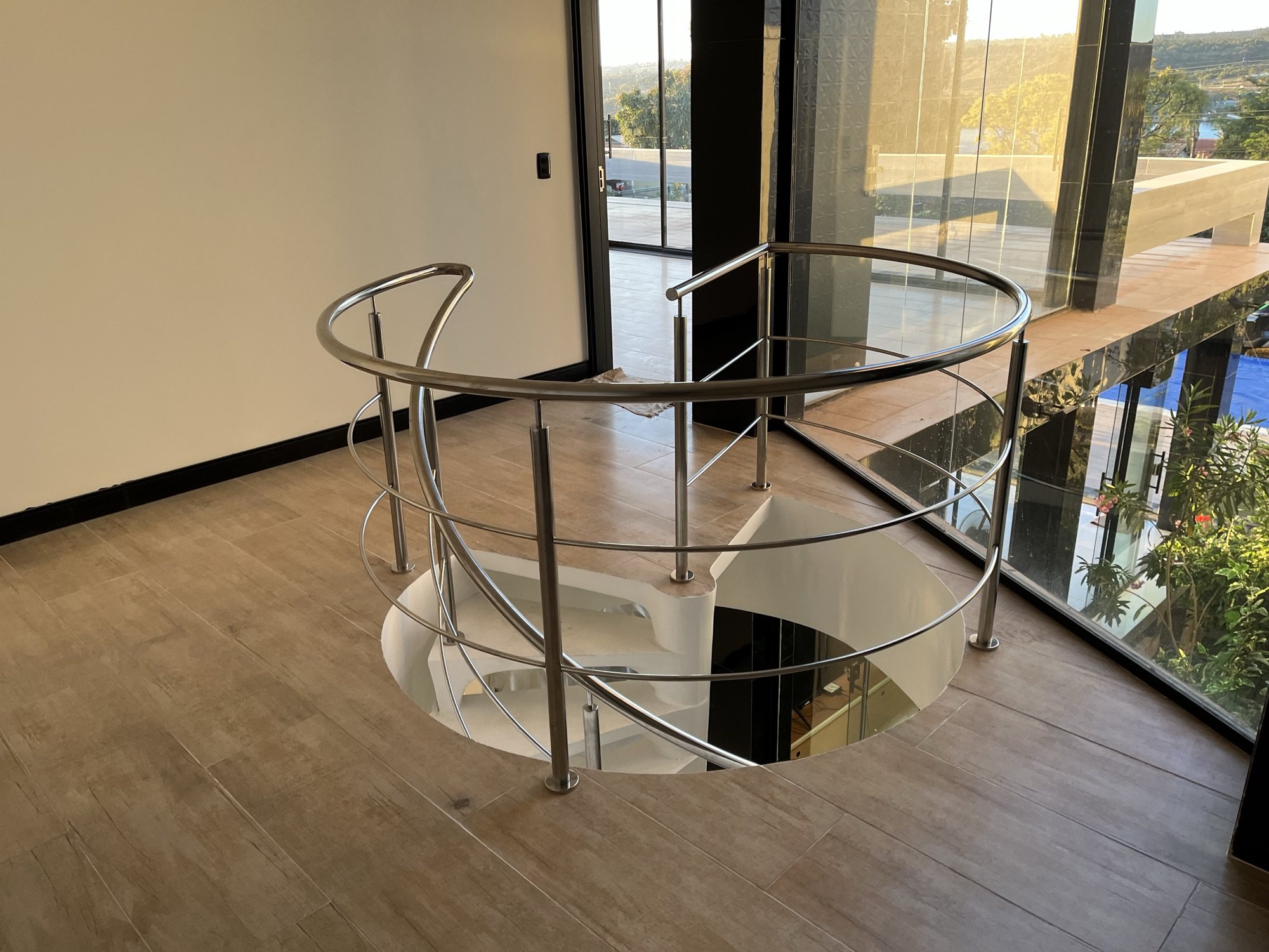 Guarda-corpo circular em aço inox ao redor da abertura de uma escada helicoidal branca, visto do pavimento superior